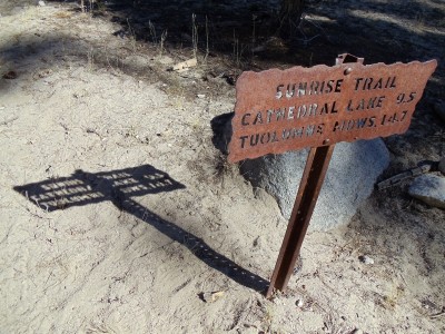 A Yosemite backcountry trail sign, marking Sunrise Trail, made of rusted metal with lettering cut through it, followed by a long midafternoon shadow, in which the gaps of the sign's letters are visible