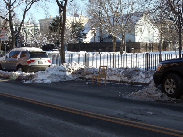 A parking space cleared of snow lies empty but for a wooden chair placed in the center of it, laying claim to that space for the person who cleared it