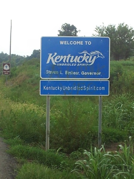 A welcome-to-Kentucky sign with spray paint graffiti across it